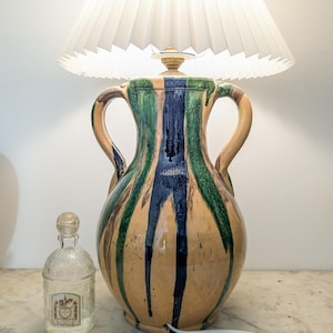 May include: A table lamp with a white pleated lampshade and a ceramic base with green, blue, and brown stripes. A small clear glass bottle with a label is on the table.