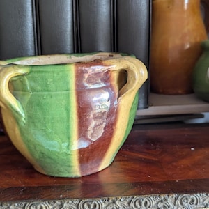 Könnte beinhalten: Ein grüner, brauner und gelber gestreifter Keramiktopf mit zwei Henkeln. Der Topf ist aus glasierter Keramik und hat einen rustikalen, Vintage-Look.