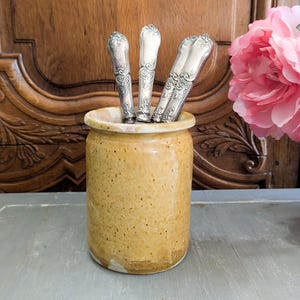 Puede incluir: Un juego de cuatro utensilios plateados con mangos ornamentados se muestran en un portautensilios de cerámica amarilla. El portautensilios tiene un esmalte moteado y está sentado sobre una superficie gris.