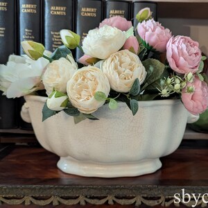 Puede incluir: Un tazón de cerámica blanco con un esmalte craquelado lleno de un ramo de flores artificiales blancas y rosas. Las flores están dispuestas en un racimo con hojas y tallos verdes.