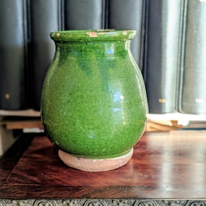 May include: A small, round, green ceramic vase with a textured surface. The vase sits on a light brown base and has a slightly flared rim. The background features a row of dark green books.