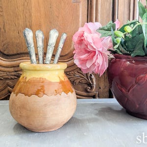 Puede incluir: Una maceta de terracota con un esmalte amarillo contiene un grupo de cubiertos de plata. La maceta está sentada sobre una superficie gris frente a un armario de madera. Una maceta de terracota roja con un diseño floral contiene flores rosas.