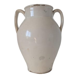 May include: Cream-colored ceramic vase with two handles. The vase has a wide body that tapers towards the base and a slightly flared rim. The surface has a glossy finish with some imperfections, giving it a rustic appearance.
