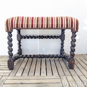 Pode incluir: Banqueta antiga com almofada listrada em vermelho, branco e dourado. A estrutura de madeira escura apresenta pernas e suportes esculpidos em espiral. A banqueta é uma peça de mobiliário decorativa, com um design clássico.