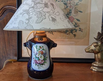 May include: A blue and white porcelain lamp base with floral designs and gold accents. The lamp has a white lampshade with a gray lace pattern.