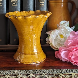 May include: A yellow ceramic vase with a scalloped rim. The vase is sitting on a wooden surface with a decorative trim.