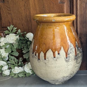 Op de afbeelding: Een grote keramische pot in antieke stijl met een brede opening. De pot heeft een tweekleurig ontwerp, met een lichtbeige basis en een druipende, glanzende bruine glazuur aan de bovenkant. De pot staat tegen een houten achtergrond.