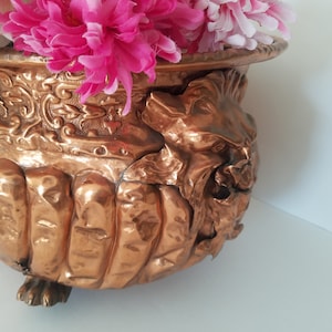 Peut inclure: Un pot en cuivre avec un design décoratif représentant une tête de lion et des pieds. Le pot est rempli de fleurs roses et blanches.