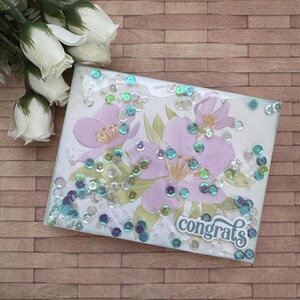 May include: A handmade card with a floral design and the word "congrats" in a decorative font. The card is decorated with iridescent sequins.