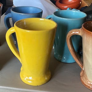 May include: A collection of ceramic mugs in various colors including bright yellow, mint green, light blue, orange, and a tan mug with brown rim detailing. Each mug features a curved handle and a flared base, arranged together in a group.