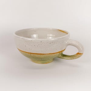 May include: A handmade ceramic mug with a speckled off-white body, a green base, and a yellow-orange stripe. The mug has a rounded handle and is suitable for coffee, tea, or other beverages.