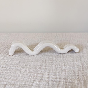 May include: A white ceramic chopstick rest in a wavy, serpentine design. The rest has a glossy finish with small, dark speckles. The object is placed on a textured, light-colored surface.