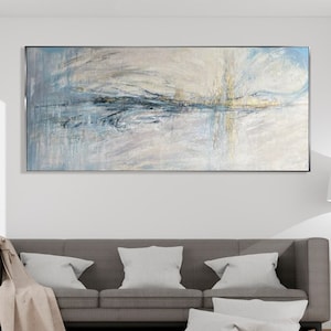 Puede incluir: Un salón moderno con un sofá gris, cojines blancos y una manta beige. Encima del sofá cuelga una gran pintura abstracta en tonos azules, blancos y grises. Una lámpara de pie trípode de madera y una planta en maceta completan la decoración.