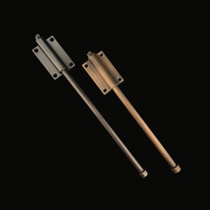 Könnte beinhalten: Zwei Türriegel aus Metall. Einer ist dunkelgrau, der andere messingfarben. Jeder Riegel hat eine rechteckige Montageplatte mit vier Schraubenlöchern und eine lange, schlanke Stange, die sich davon erstreckt.