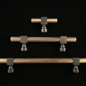 May include: Three gold cabinet pulls with black square accents and round bases. The pulls are arranged in a triangular pattern, with the smallest pull at the top and the largest pull at the bottom.
