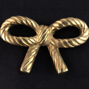 Op de afbeelding: Een gouden metalen strik met een touwachtige textuur. De strik is in een knoop geknoopt en heeft een gladde, gepolijste afwerking.