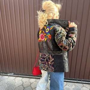 May include: A black jacket with colourful embroidery on the back. The jacket is made of a patchwork of different fabrics and has a collar. The person wearing the jacket is also wearing blue jeans and red shoes.