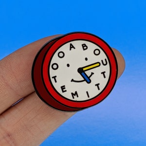 May include: Red enamel pin shaped like a clock face with the words "ABOUT TIME" written around the edge. The clock hands are pointing to the 12 and the 3, forming a smiley face.