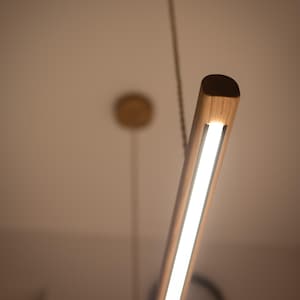 Può includere: Una moderna lampada a sospensione in legno di forma rettangolare. La luce è emessa da una striscia LED bianca e brillante, incastonata in legno chiaro. La lampada è sospesa al soffitto tramite un sottile filo.