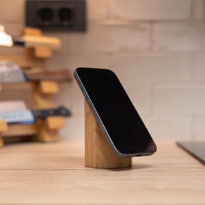 May include: A black smartphone rests on a wooden phone stand. The stand is made of light-colored wood and has a cylindrical base. The phone is angled slightly, displaying a black screen. The background is blurred.