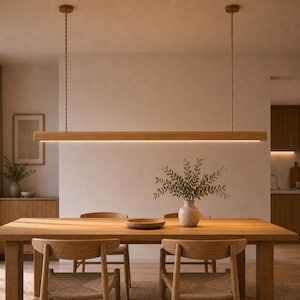 Puede incluir: Una lámpara colgante lineal que presenta una viga de madera horizontal larga con una tira de luz LED integrada, suspendida del techo por dos cables trenzados delgados. La luz emite un brillo cálido hacia abajo sobre una mesa de madera.