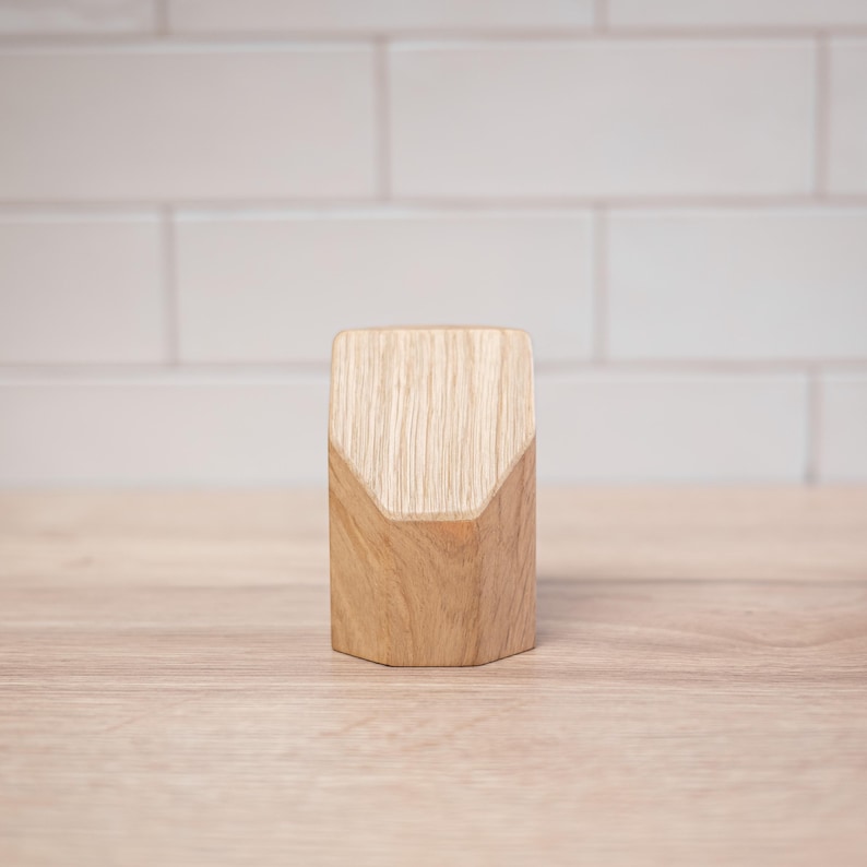 May include: A wooden bottle stopper with a geometric design. The stopper is made of light-colored wood with a natural grain pattern. The top of the stopper has a unique angled cut, adding a modern touch. It stands on a light-colored wooden surface.