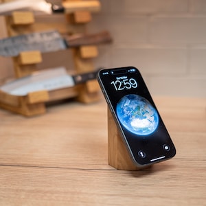 May include: A smartphone displaying the time 12:59 and a blue Earth image, resting on a small, angled wooden stand. The stand is made of light-colored wood. A wooden knife block with several knives is in the background.