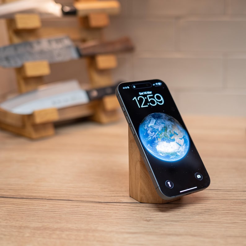 May include: A smartphone displaying the time 12:59 and a blue Earth image, resting on a small, angled wooden stand. The stand is made of light-colored wood. A wooden knife block with several knives is in the background.