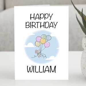 Puede incluir: Tarjeta de cumpleaños blanca con el texto negro "HAPPY BIRTHDAY WILLIAM". La tarjeta presenta una ilustración de un ratón sosteniendo globos de colores sobre un fondo de cielo azul claro.