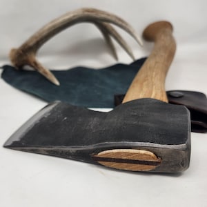 Puede incluir: Un hacha de estilo vintage con mango de madera y cabeza metálica oscura. La imagen también incluye una funda de cuero y cuernos de ciervo. El conjunto evoca un tema rústico y campestre.