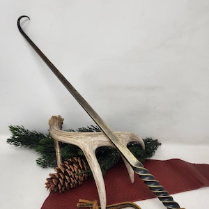 Pode incluir: Um atiçador de lareira artesanal, de cor bronze, com uma pega torcida e uma extremidade curva. O atiçador é exibido com uma chifre de veado, agulhas de pinheiro, uma pinha e um pedaço de couro vermelho.