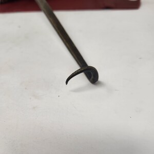 May include: A dark metal tool with a curved end and a long, straight handle. The tool is likely used for prying or lifting. The background includes a red surface and some greenery.