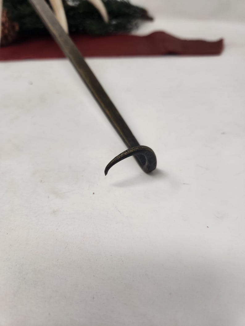 May include: A dark metal tool with a curved end and a long, straight handle. The tool is likely used for prying or lifting. The background includes a red surface and some greenery.