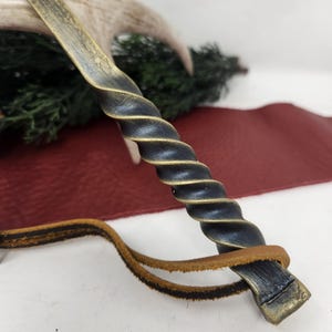 May include: A handcrafted, twisted metal wand with a dark blue and gold finish. The wand features a spiral design on the handle and a leather strap. The background includes a deer antler and greenery.