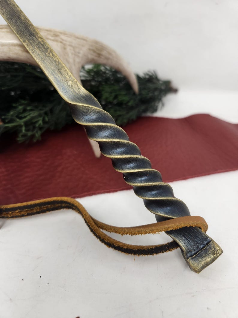 May include: A handcrafted, twisted metal wand with a dark blue and gold finish. The wand features a spiral design on the handle and a leather strap. The background includes a deer antler and greenery.