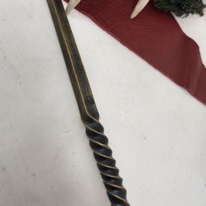 May include: A handcrafted wand with a dark, textured finish and a spiraled handle. The wand has a leather strap attached to the end. The wand is lying on a white surface with a red leather strip, antlers, and greenery in the background.