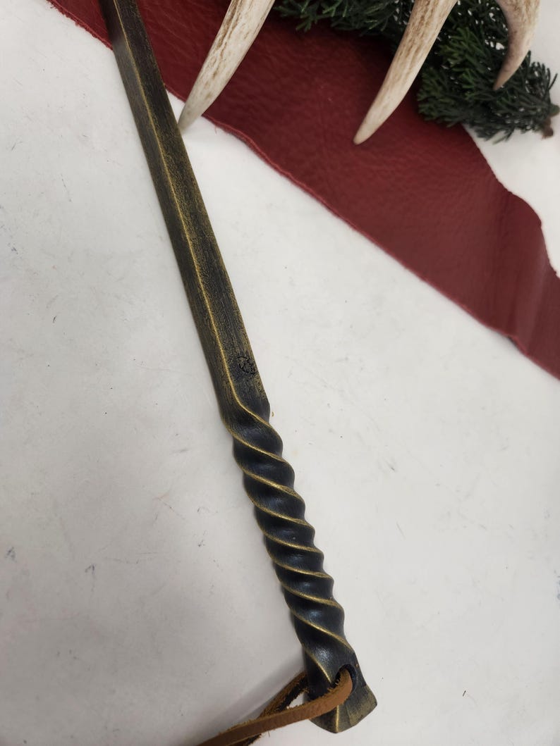 May include: A handcrafted wand with a dark, textured finish and a spiraled handle. The wand has a leather strap attached to the end. The wand is lying on a white surface with a red leather strip, antlers, and greenery in the background.