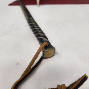 May include: A hand-forged, black metal fire poker with a twisted shaft and a brass head. A brown leather loop is attached to the head. The poker is designed for use with a fireplace or campfire.