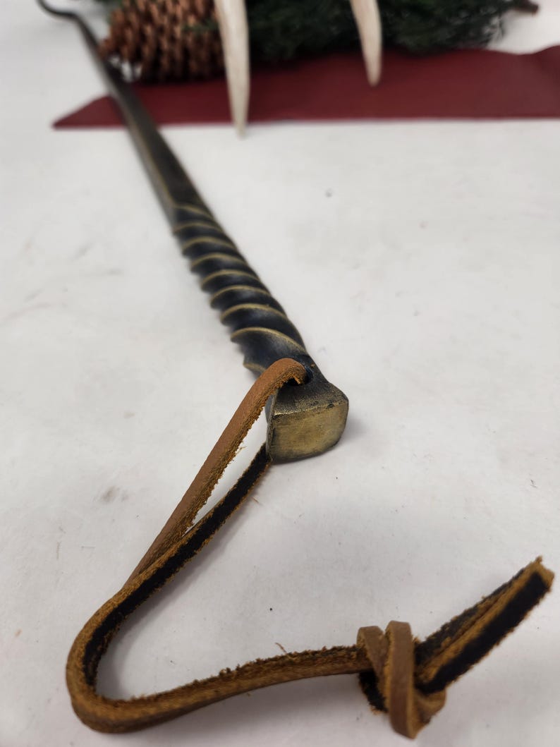 May include: A hand-forged, black metal fire poker with a twisted shaft and a brass head. A brown leather loop is attached to the head. The poker is designed for use with a fireplace or campfire.