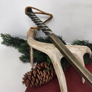 May include: A bronze-colored letter opener with a spiraled handle and a leather loop. The opener rests on a deer antler, with a pine cone and evergreen sprigs in the background. The opener has a rustic, antique aesthetic.