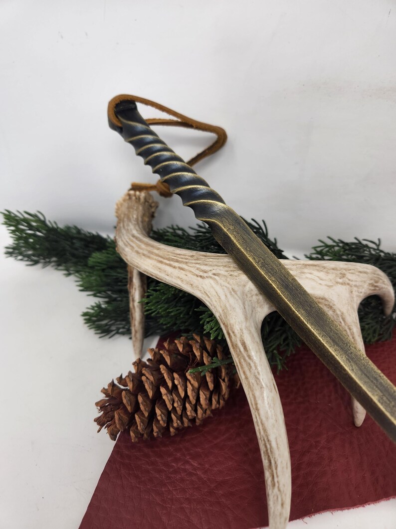 May include: A bronze-colored letter opener with a spiraled handle and a leather loop. The opener rests on a deer antler, with a pine cone and evergreen sprigs in the background. The opener has a rustic, antique aesthetic.