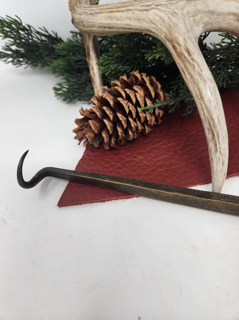 May include: A rustic still life featuring a brown pine cone, a curved metal tool, and a section of a deer antler. The objects rest on a piece of red leather and are set against a backdrop of green foliage.