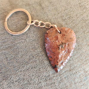 May include: A heart-shaped keychain pendant made of reddish-brown stone, attached to a silver-colored chain and keyring. The stone has a textured surface with darker spots and a pointed bottom. The background is a textured, neutral-toned surface.