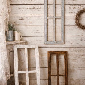 May include: Three rectangular wooden window frames with a multi-pane design. One frame is mounted on the wall with a weathered light blue finish, while two others stand on the floor, one in a distressed white finish and one in a dark brown wood stain.