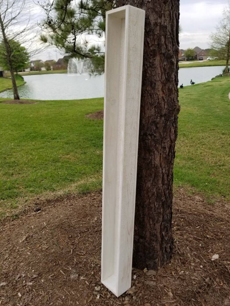 May include: A white wooden planter box with a rectangular shape. The planter is standing upright against a tree trunk.