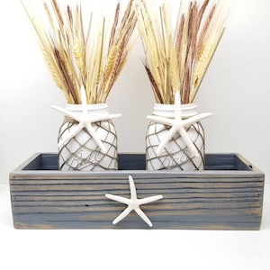 May include: Two white mason jars with wheat and starfish decorations sit in a gray wooden tray. The jars are wrapped in netting and adorned with white starfish. The tray also features a starfish. A rustic, beach-themed decorative piece.