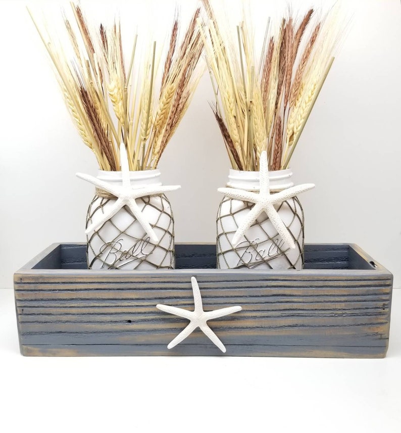 May include: Two white mason jars with wheat and starfish decorations sit in a gray wooden tray. The jars are wrapped in netting and adorned with white starfish. The tray also features a starfish. A rustic, beach-themed decorative piece.