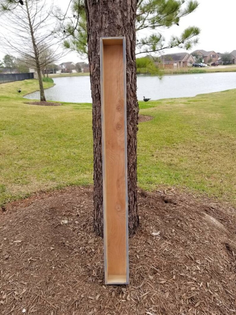 May include: A long, narrow wooden planter box with a light blue painted edge. The planter is leaning against a tree trunk in a grassy area.