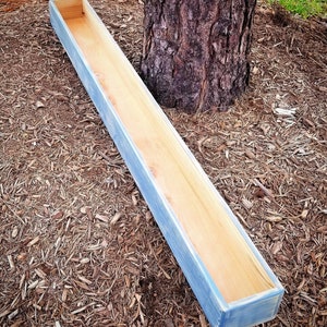 May include: A long, rectangular wooden planter box with a light blue wash finish. The planter is made of natural wood and has a rustic look.