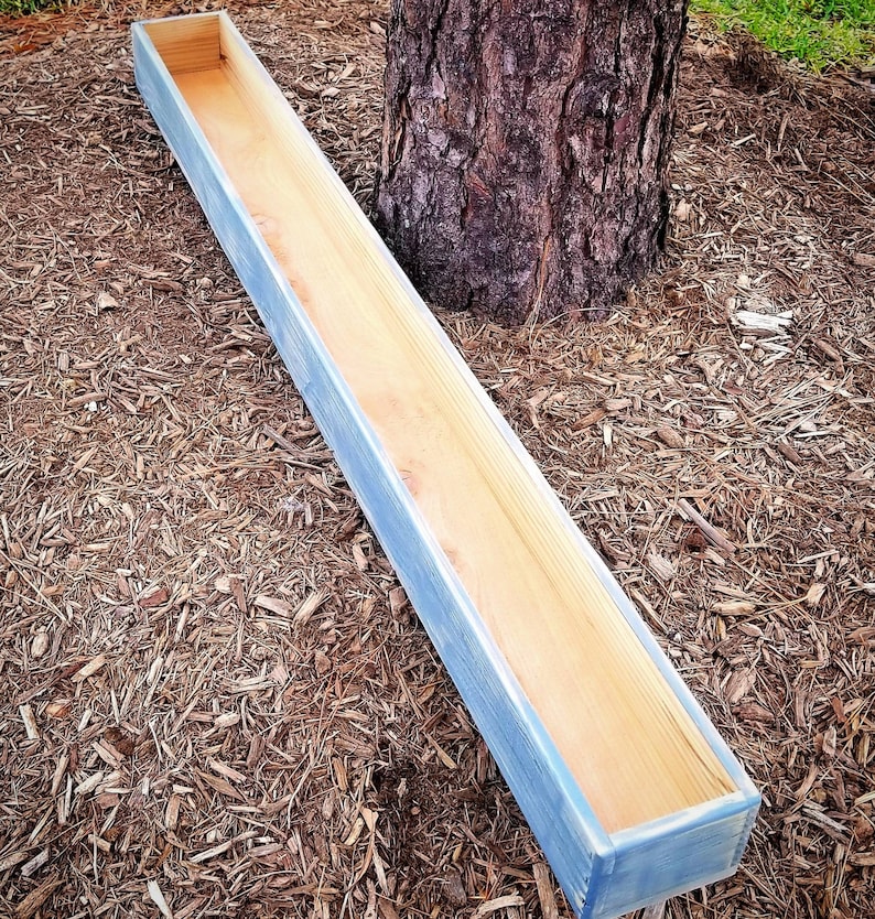 May include: A long, rectangular wooden planter box with a light blue wash finish. The planter is made of natural wood and has a rustic look.